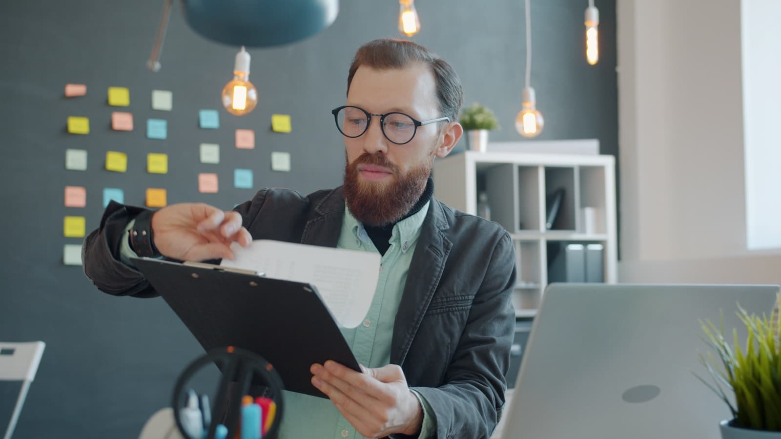 Businessman with beard and glasses reviewing documents in modern office with sticky notes on wall