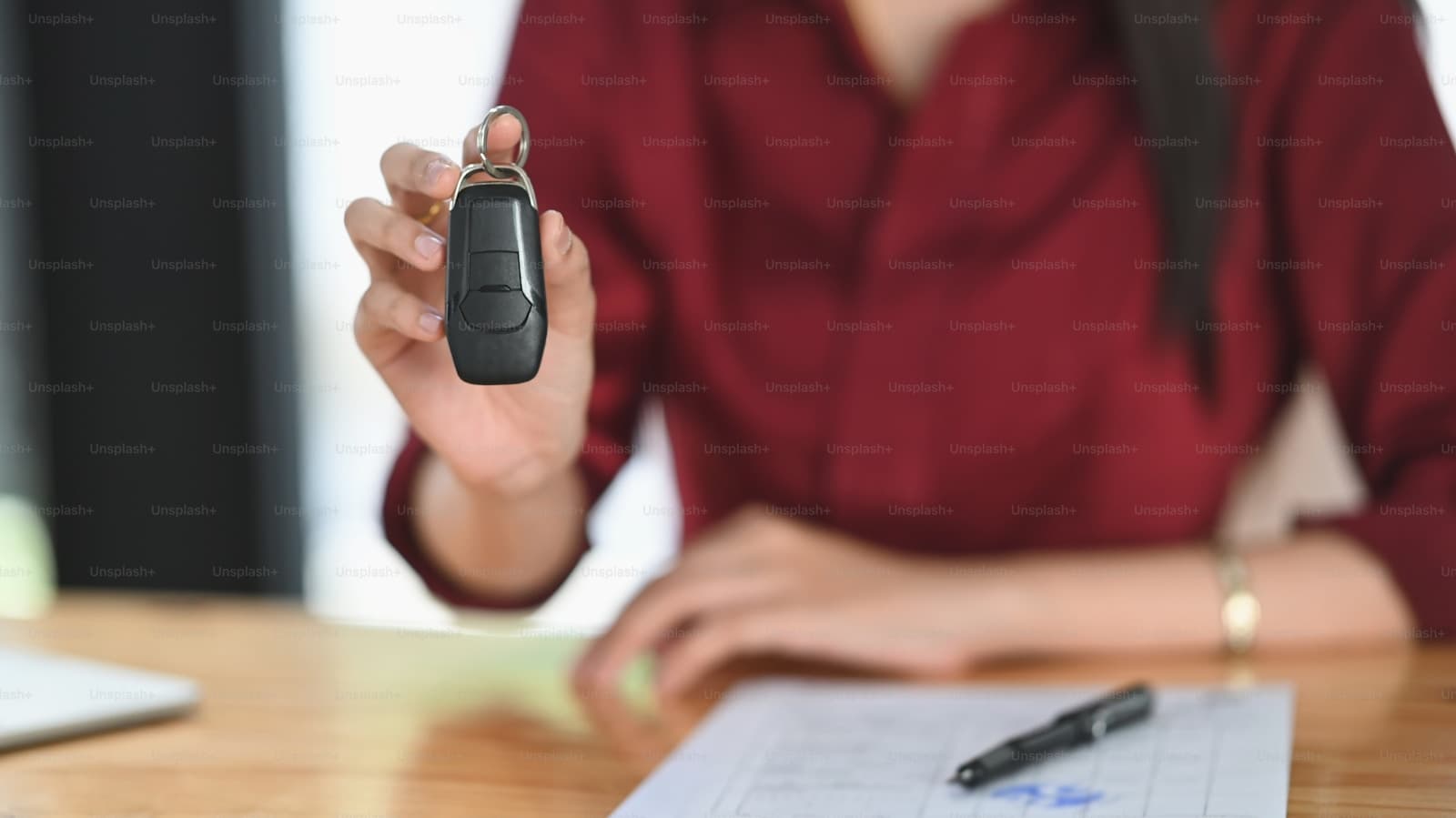 Person in red jacket holding car keys over desk with paperwork, suggesting vehicle purchase or dealership transaction