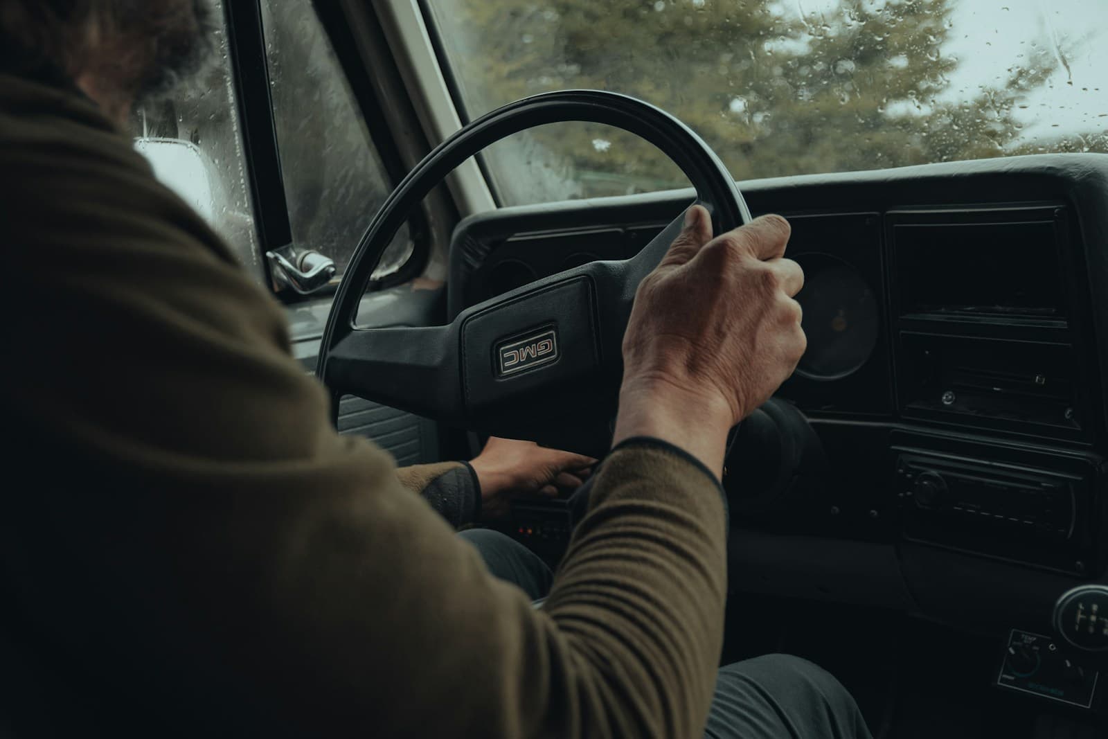 Driver in a tan shirt gripping the wheel of an older car on a tree-lined road