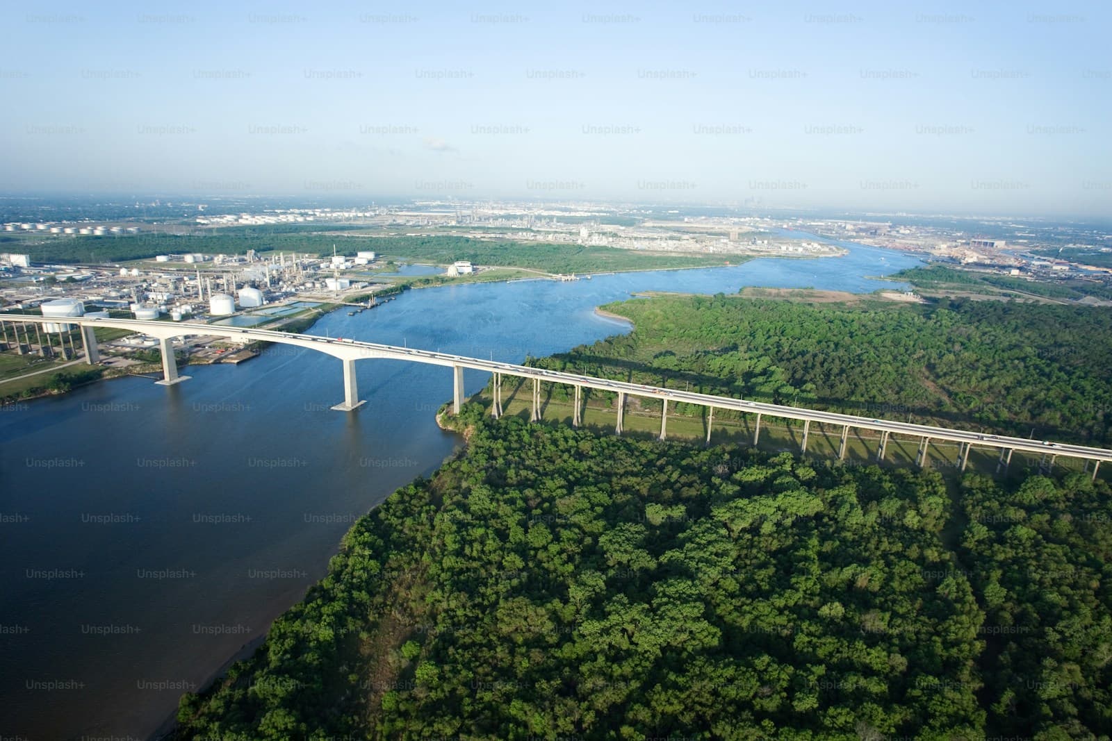 Aerial view of concrete bridge crossing river through green forested landscape with urban skyline in background