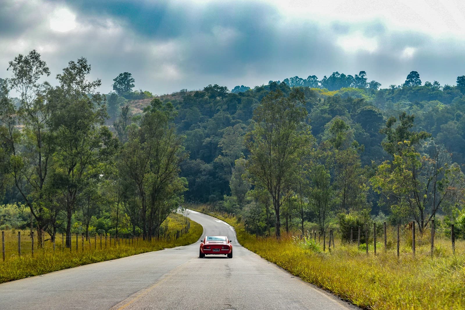 Red car driving on rural road through rolling hills with trees and cloudy sky