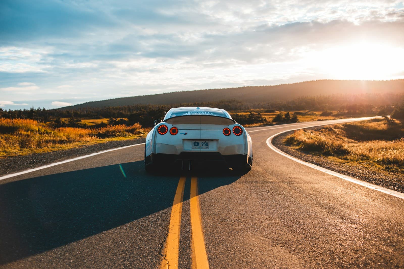 Silver sports car driving on curved rural highway during sunset with golden hills and dramatic sky