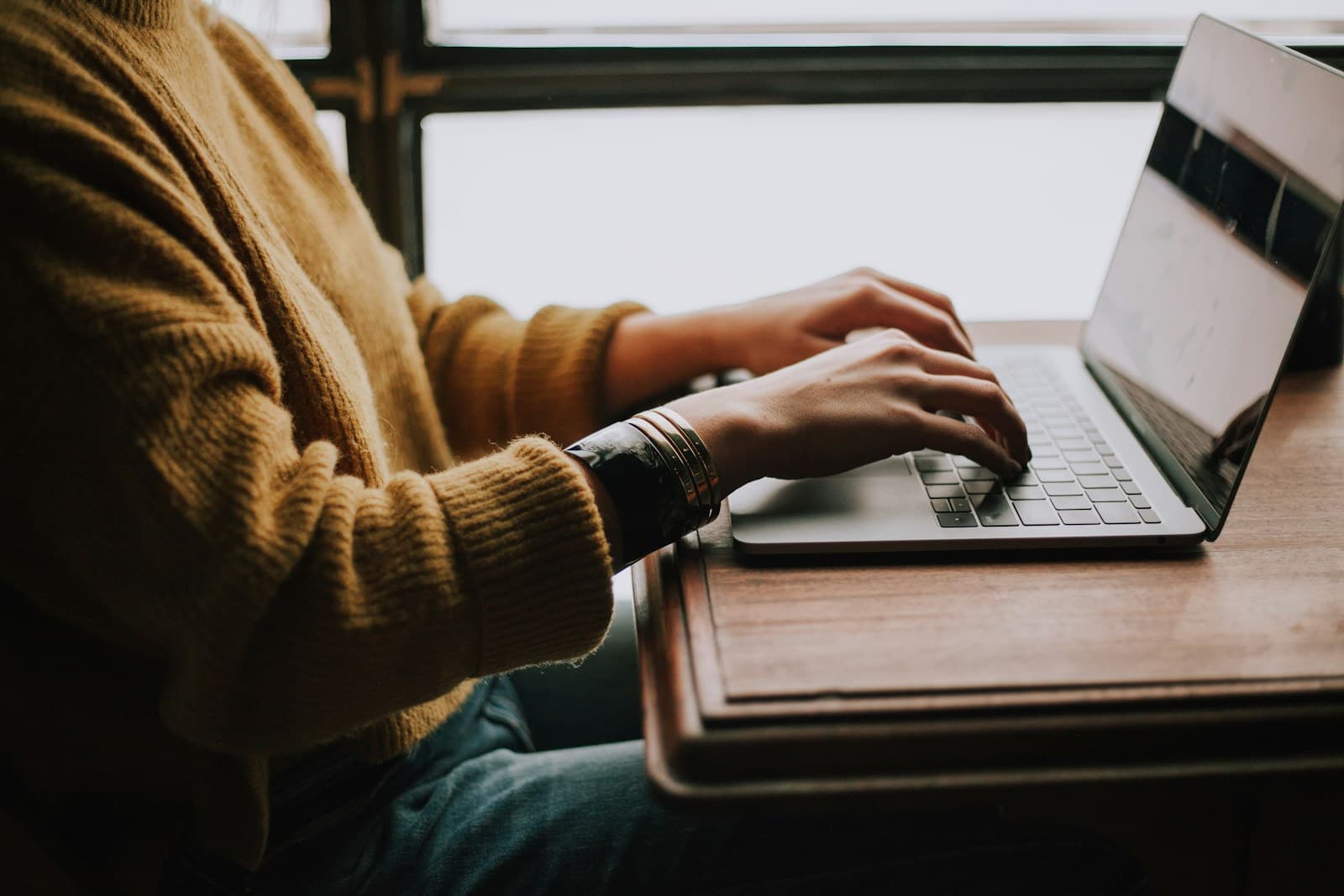Person in a mustard cardigan typing on a laptop at a desk by a window