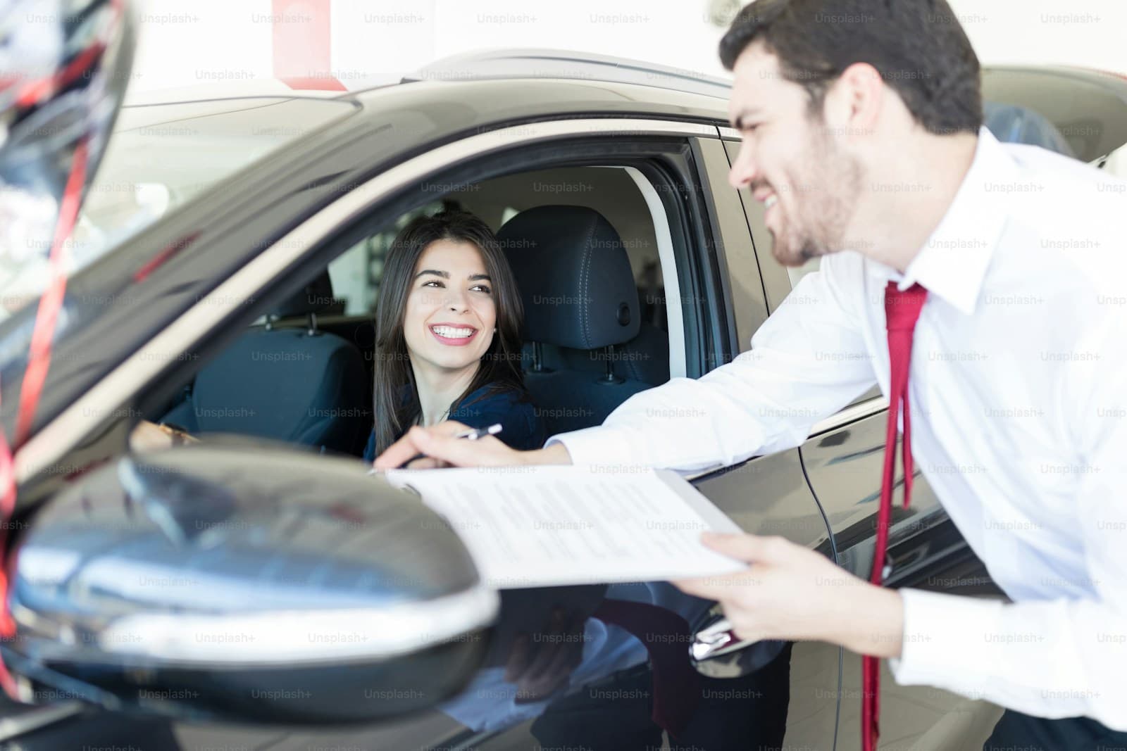 Man in a white shirt and red tie handing paperwork to a smiling woman in the driver's seat