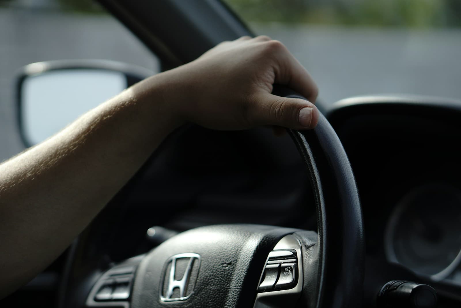Hand gripping a black Honda steering wheel with the side mirror visible while driving
