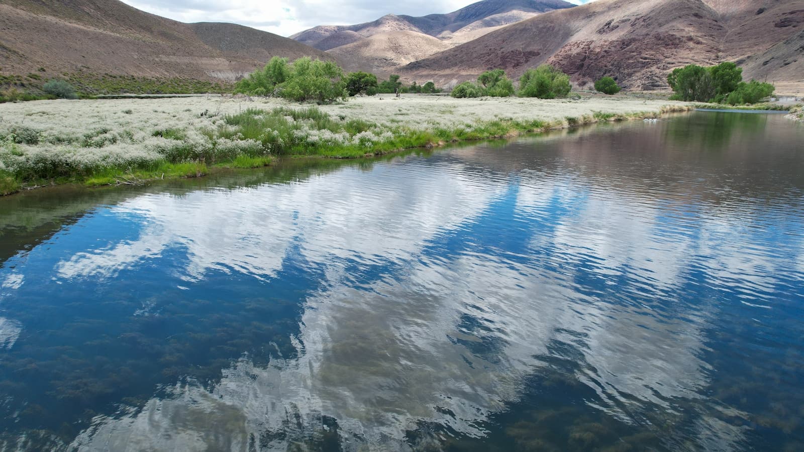 River reflecting white clouds in arid landscape with brown hills and green vegetation along the banks