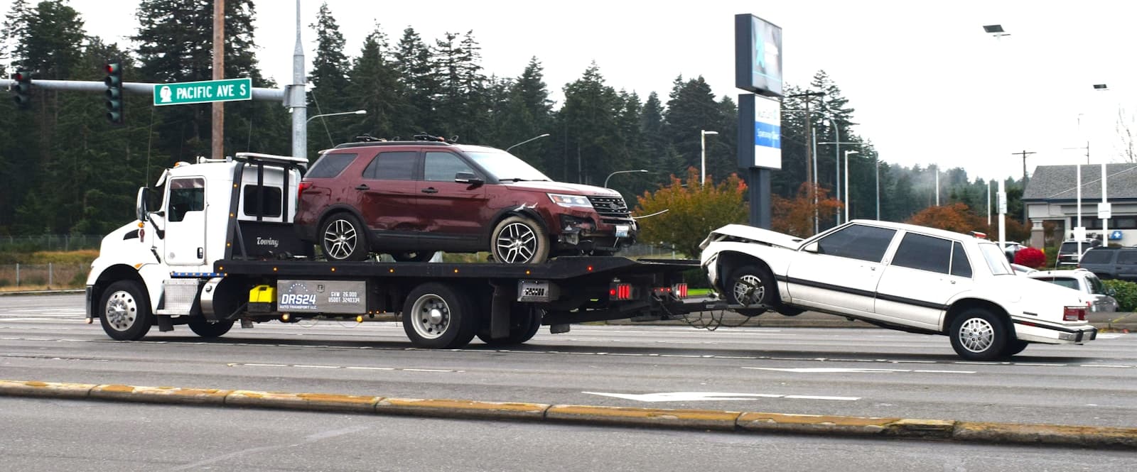 White tow truck hauling a damaged red SUV and white sedan along a daytime highway